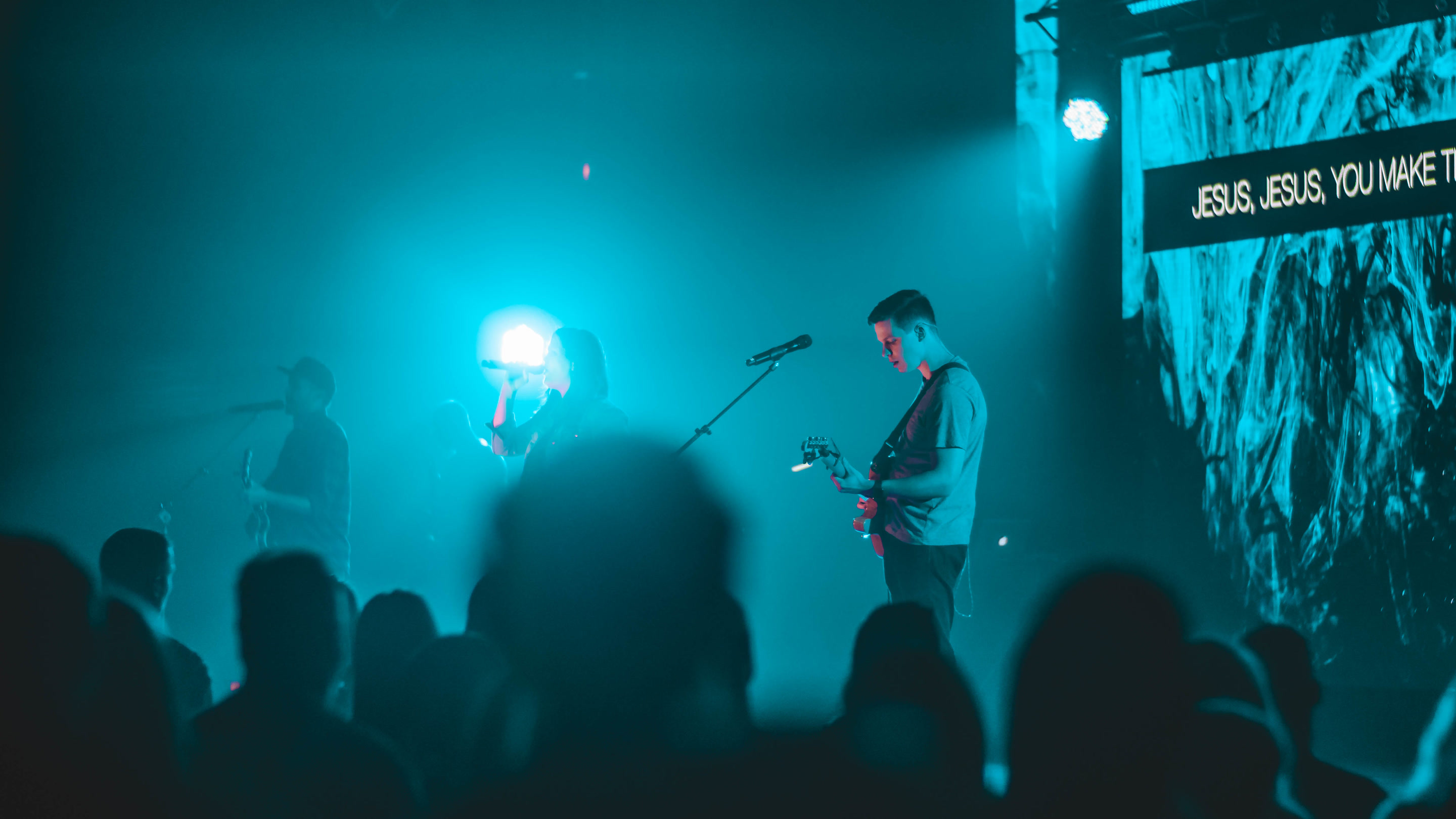 A live stage under cyan lighting, with an LED wall showing lyrics, a band, and a silhouetted audience.